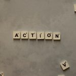 Flat lay of Scrabble tiles spelling 'Action' on a gray background, surrounded by random letters.