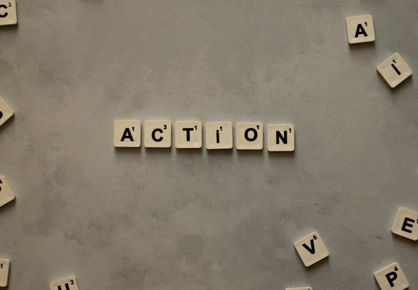 Flat lay of Scrabble tiles spelling 'Action' on a gray background, surrounded by random letters.