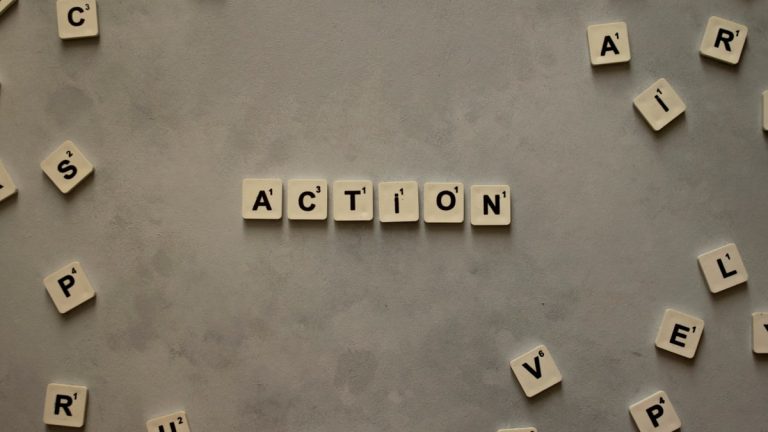 Flat lay of Scrabble tiles spelling 'Action' on a gray background, surrounded by random letters.