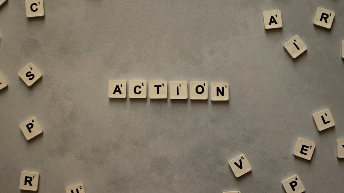 Flat lay of Scrabble tiles spelling 'Action' on a gray background, surrounded by random letters.