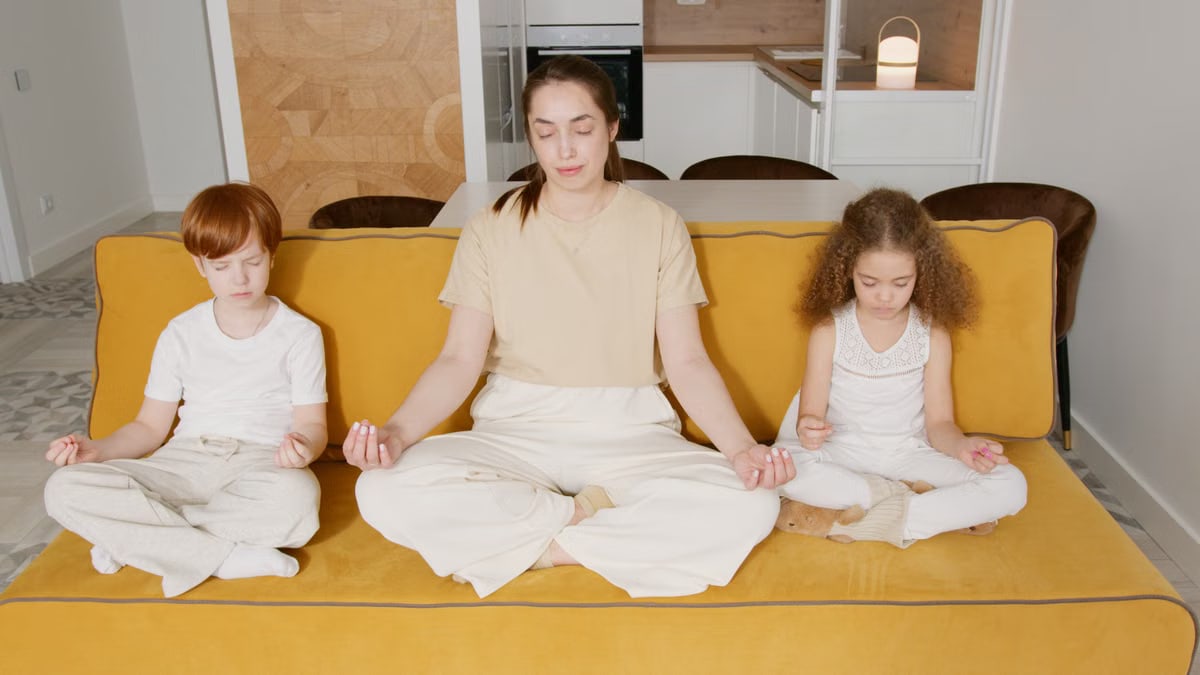 A mother and two children practicing mindfulness meditation indoors on a cozy sofa.